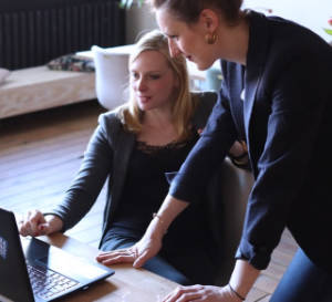 Two people in business attire. One is sitting in front of a notebook computer, while the other stands leaning forward on the desk.