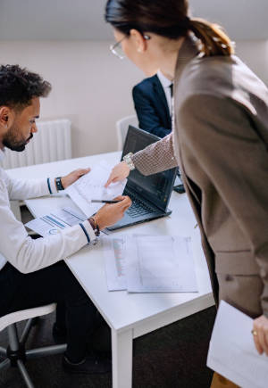 Two people in business attire. One is sitting in front of a notebook computer holding a piece of paper, while the other points to it.