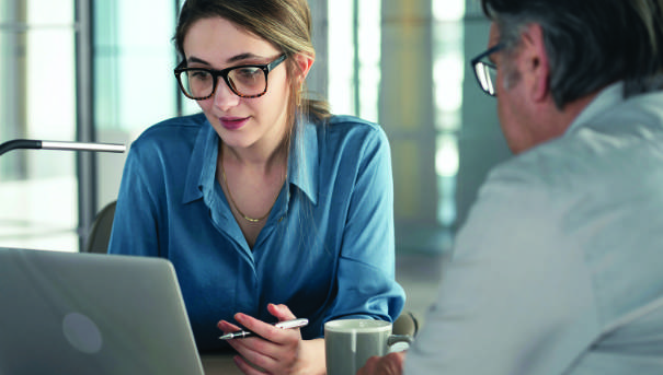 Two people in business attire sit at a table. One is holding a pen while looking at a notebook computer.